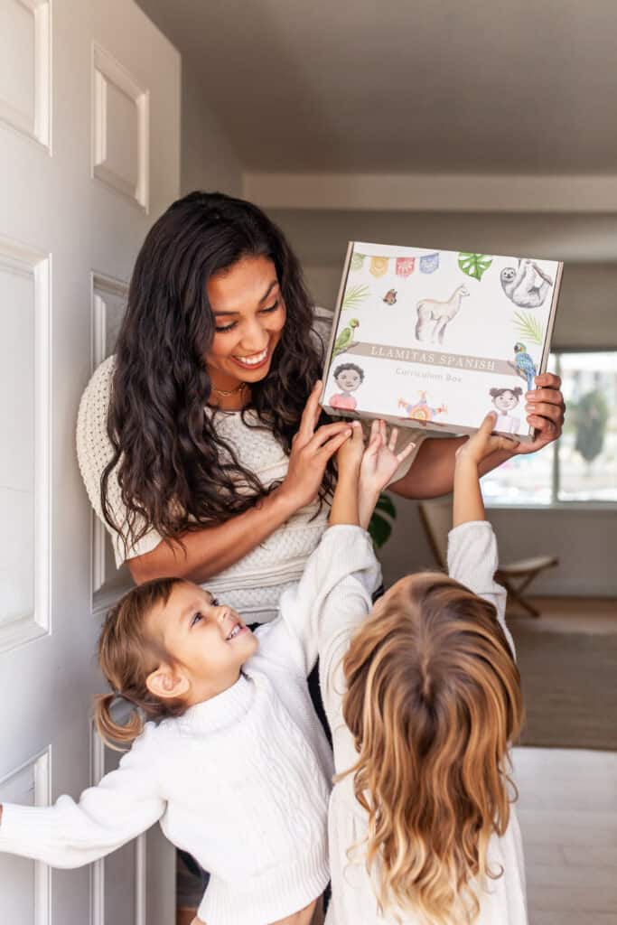 A latina mom holding up the Llamitas Spanish curriculum box at her front door and her two daughters reaching up for it.