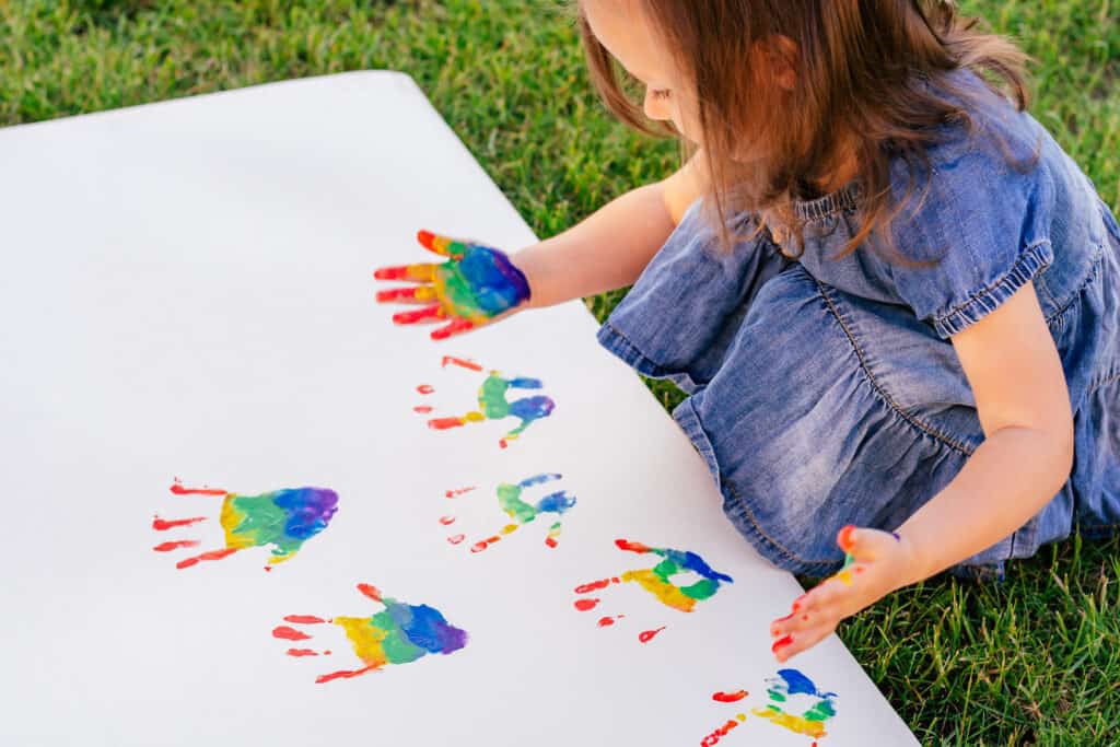 Girl with rainbow painted hands making hand prints on card
