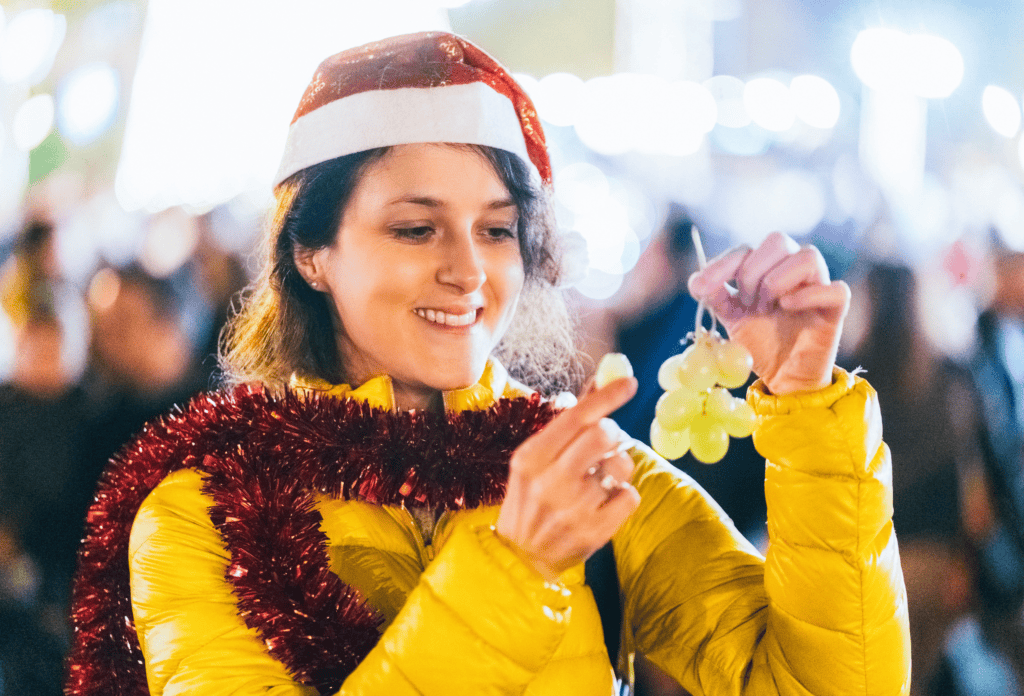 woman with santa hat on and tinsel around her neck is eating grapes counting down to midnight